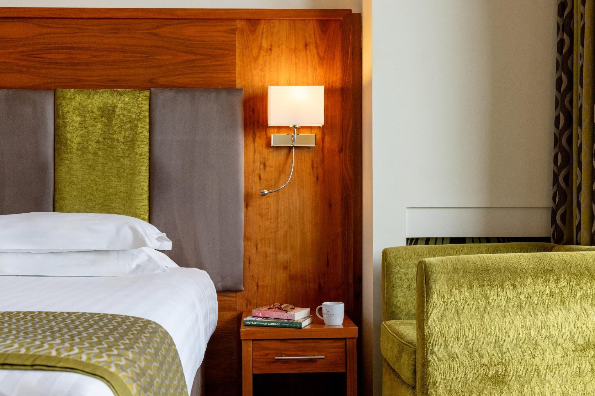 A hotel room with a wooden headboard, a bedside table with a lamp, a green armchair, and a bed with white linens.