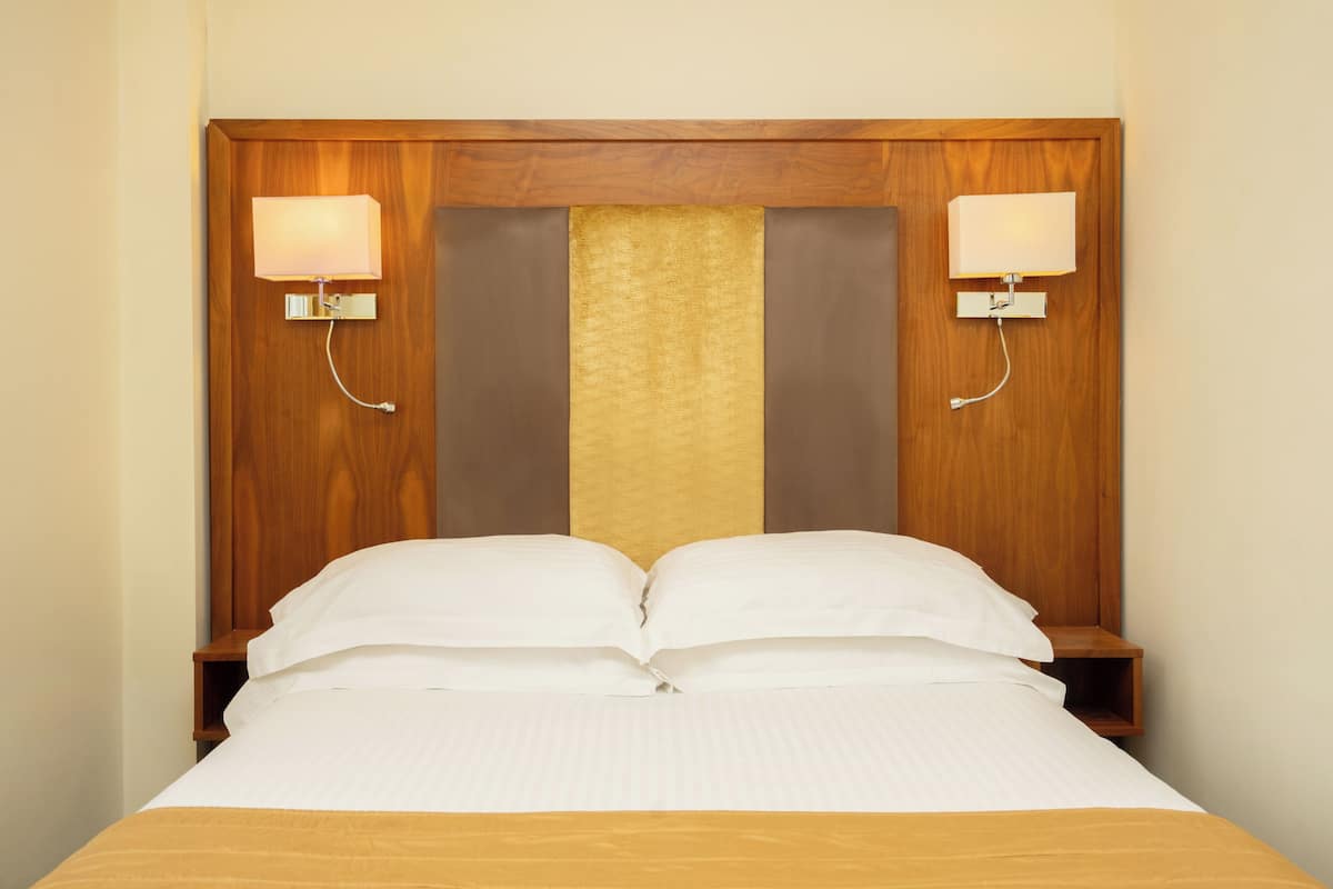 A hotel room with a wooden headboard, two bedside lamps, a bed with white bedding, and a small wooden nightstand.