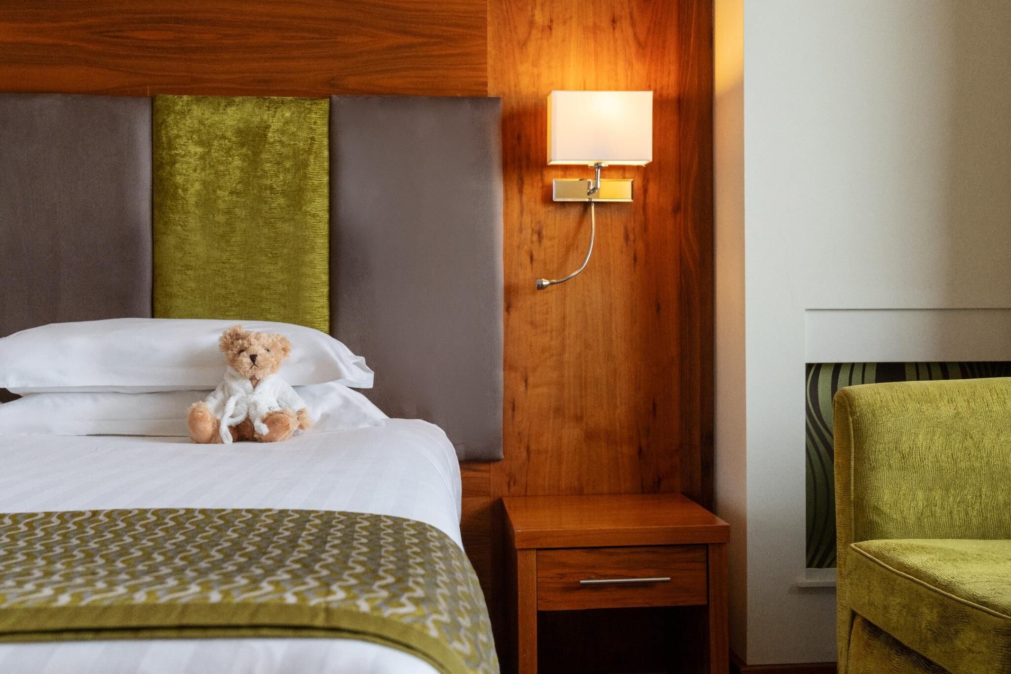 Une chambre d’hôtel avec un lit, une table de chevet, une lampe, un fauteuil vert et un mur orné d’un motif décoratif.