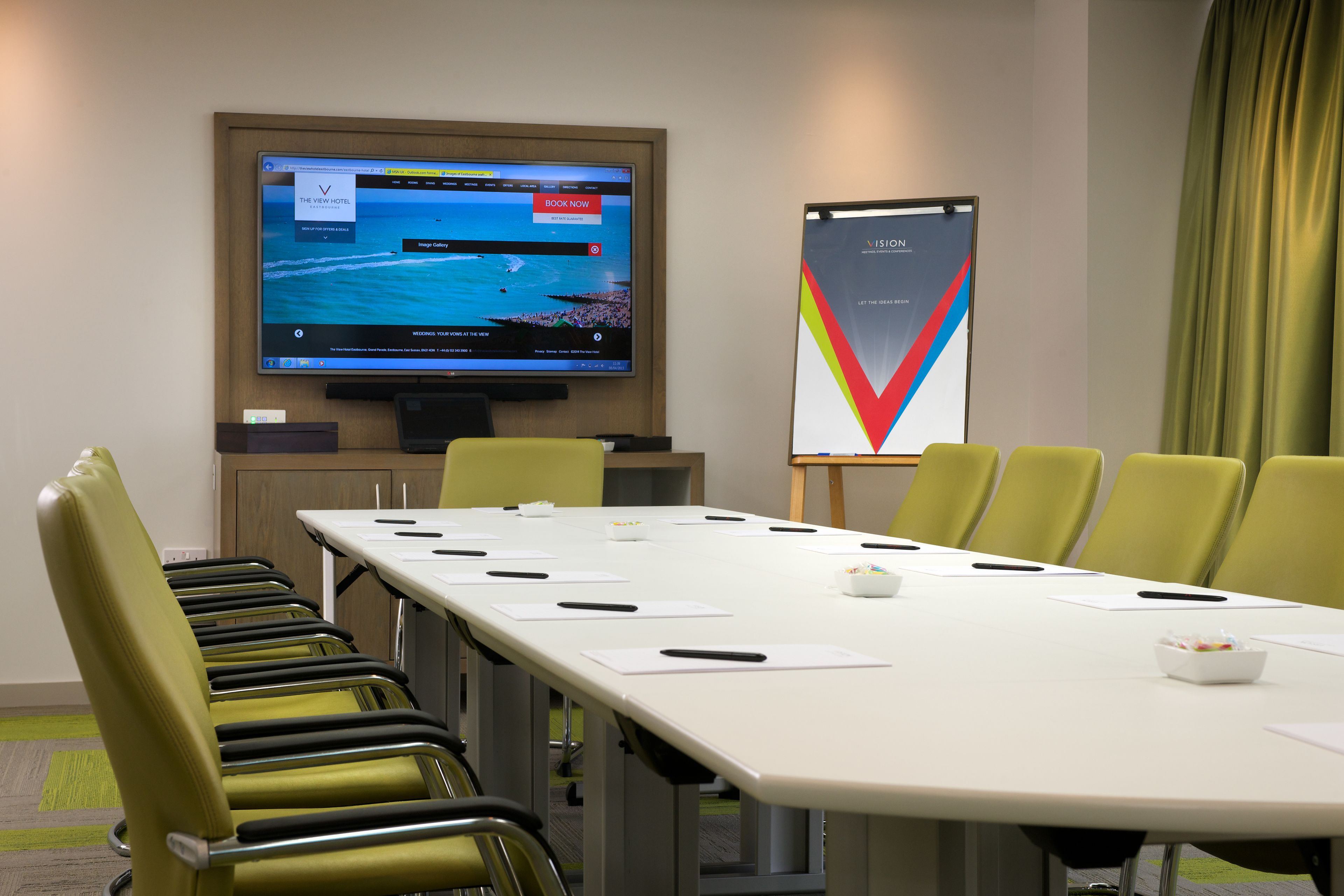 A conference room with a long table, green chairs, a screen displaying a hotel booking page, and a poster on the wall.