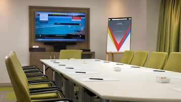 A conference room with a long table, green chairs, a screen displaying a hotel booking page, and a poster on the wall.