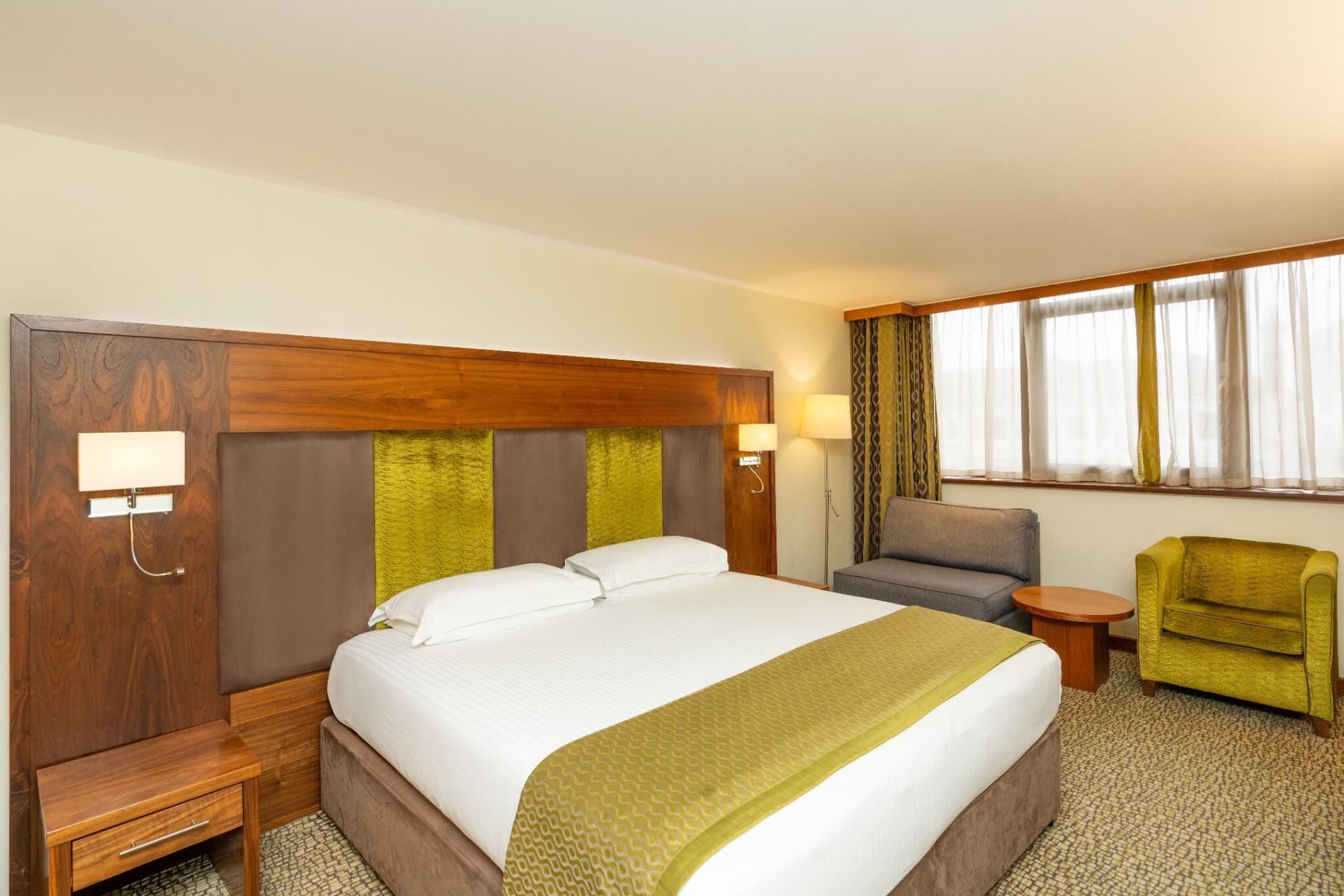 Una habitación de hotel con una cama grande, un cabecero de madera, un escritorio pequeño, una silla, una mesa redonda y una ventana con cortinas.