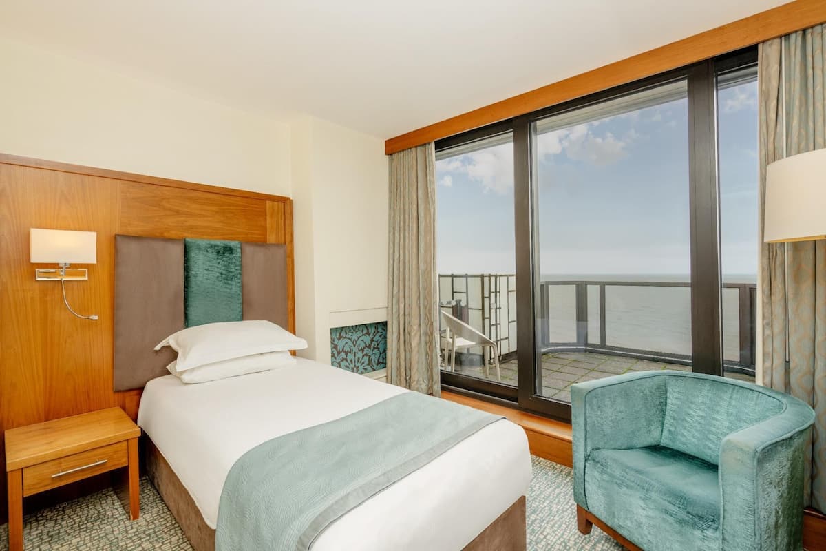 A hotel room with a bed, a nightstand, a chair, a large window with a view of the sea, and a balcony.
