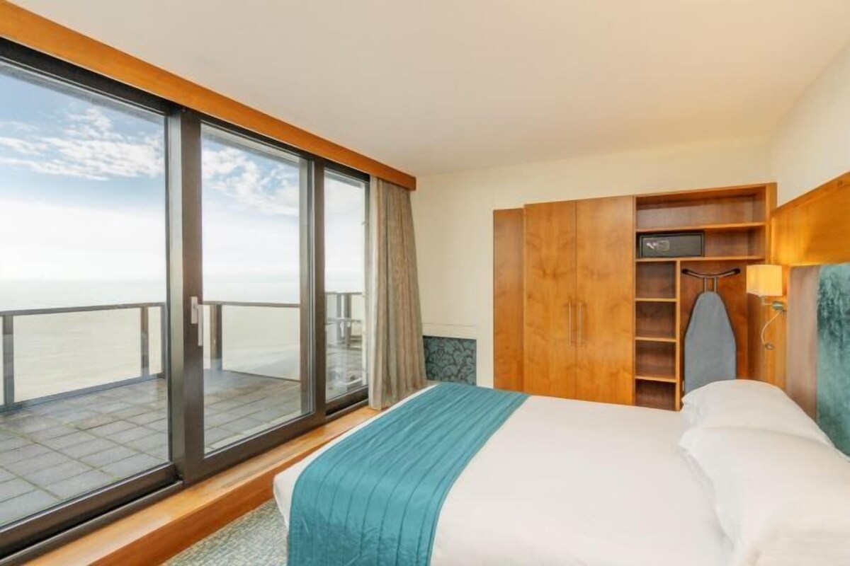 A hotel room with a large sliding glass door to the balcony, a bed with a blue accent, and wooden furniture.
