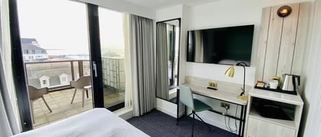 A hotel room with a large bed, a desk with a chair, a television, and a view of the city through the balcony.