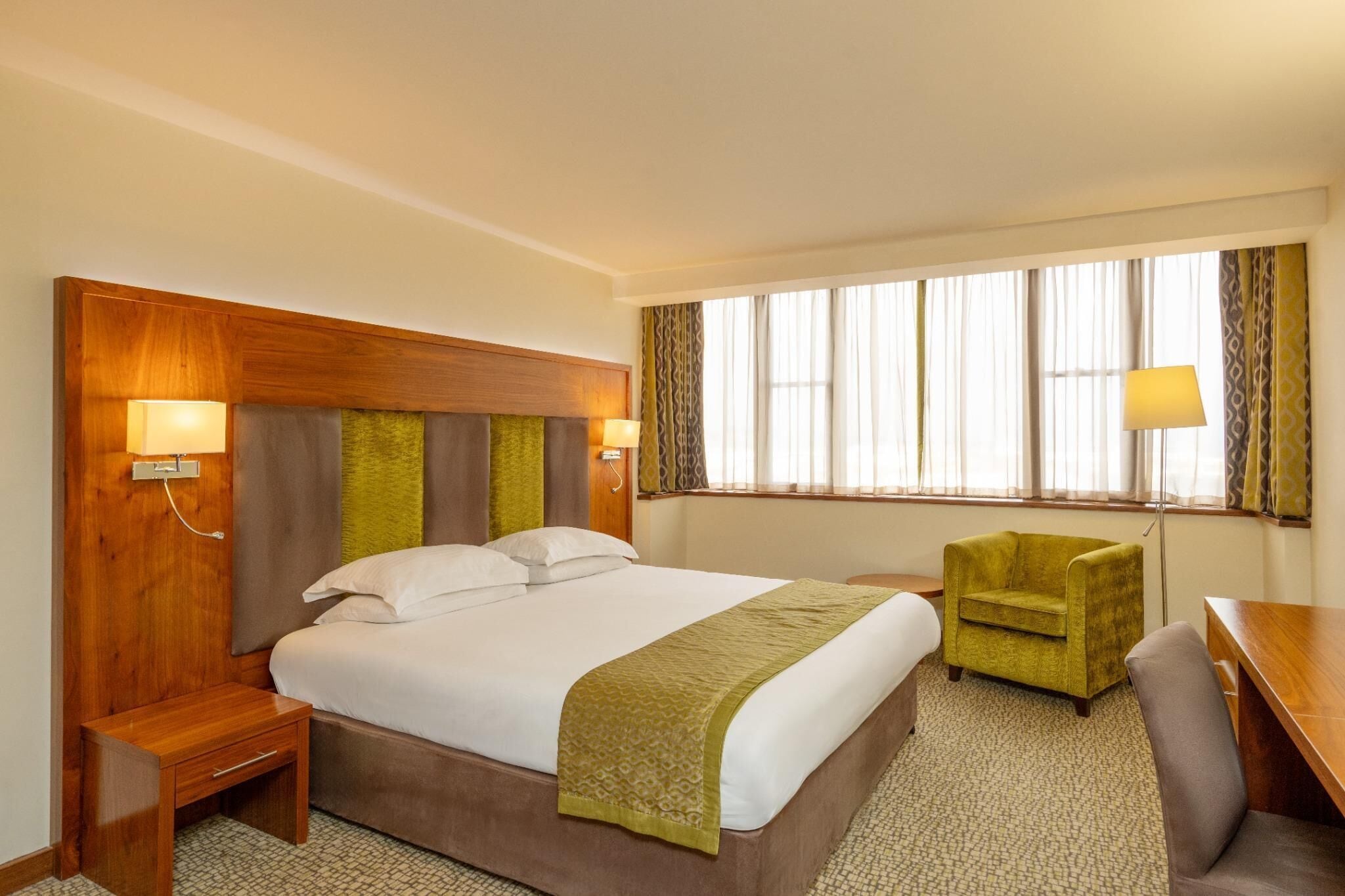 Una habitación de hotel con cama, mesitas de noche, una silla, un escritorio y una ventana con cortinas.