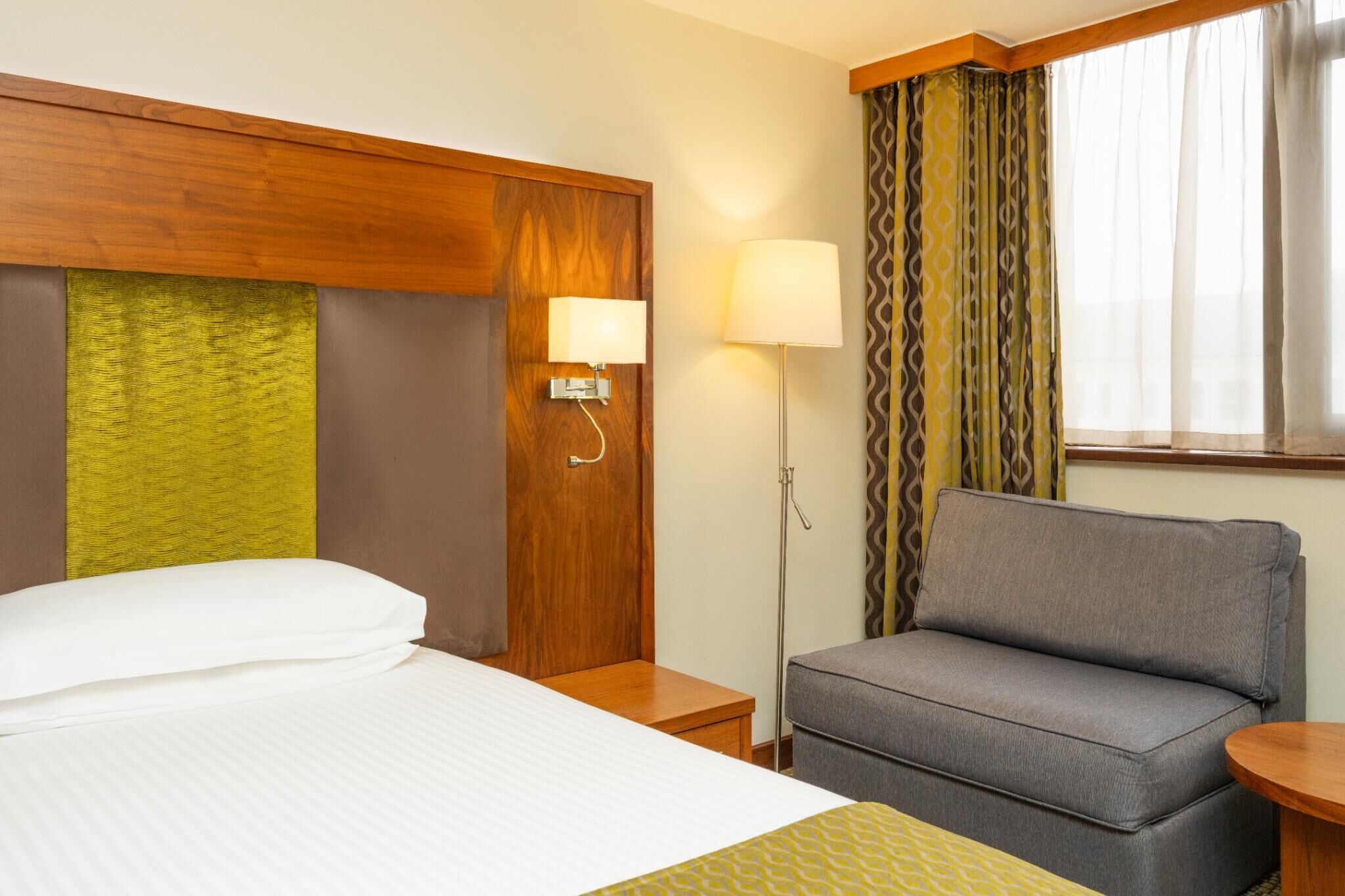 Een hotelkamer met een bed, een grijze bank, een nachtkastje, een lamp en een raam met gordijnen.
