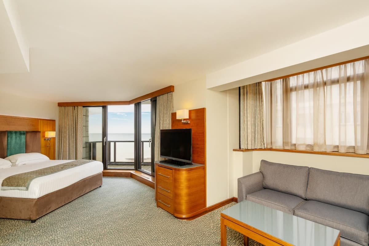 A hotel room with a bed, a sofa, a TV, a balcony with a view, and a glass table.