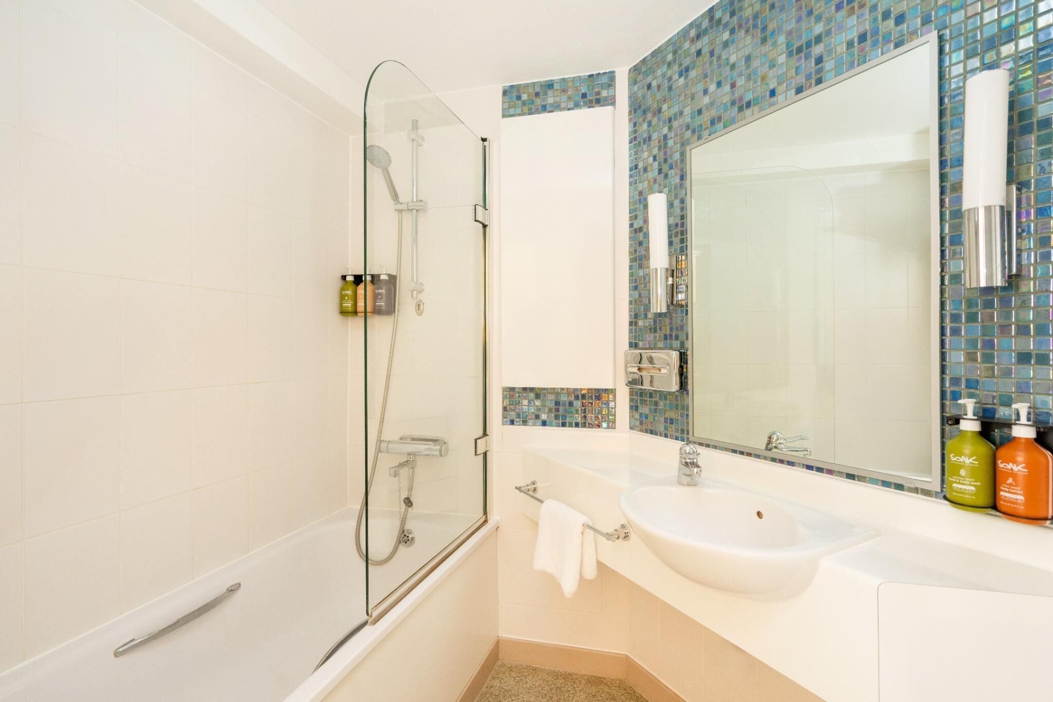 A bathroom with a glass shower enclosure, a white sink, and a mirror with a mosaic tile backsplash.