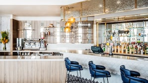 Een moderne bar met een houten toog, blauwe barkrukken en diverse flessen en glazen op planken.