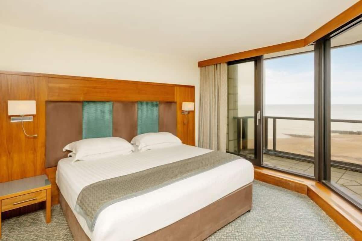 A hotel room with a bed, bedside tables, a view of the beach, and a balcony.