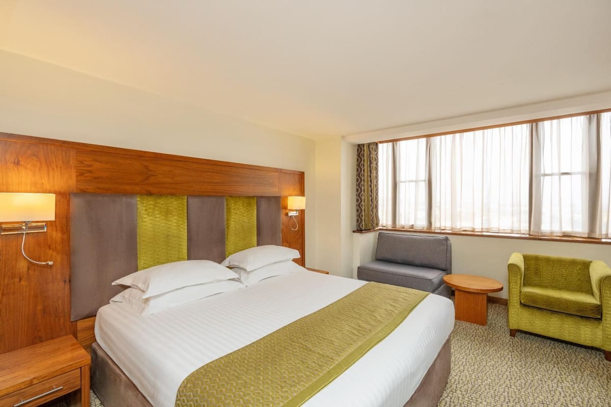 A hotel room with a large bed, a wooden headboard, a grey sofa, a green armchair, a small round table, and a window with curtains.