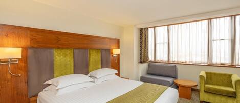 Una habitación de hotel con una cama grande, cabecera de madera, un sofá gris, un sillón verde, una mesita redonda y una ventana con cortinas.