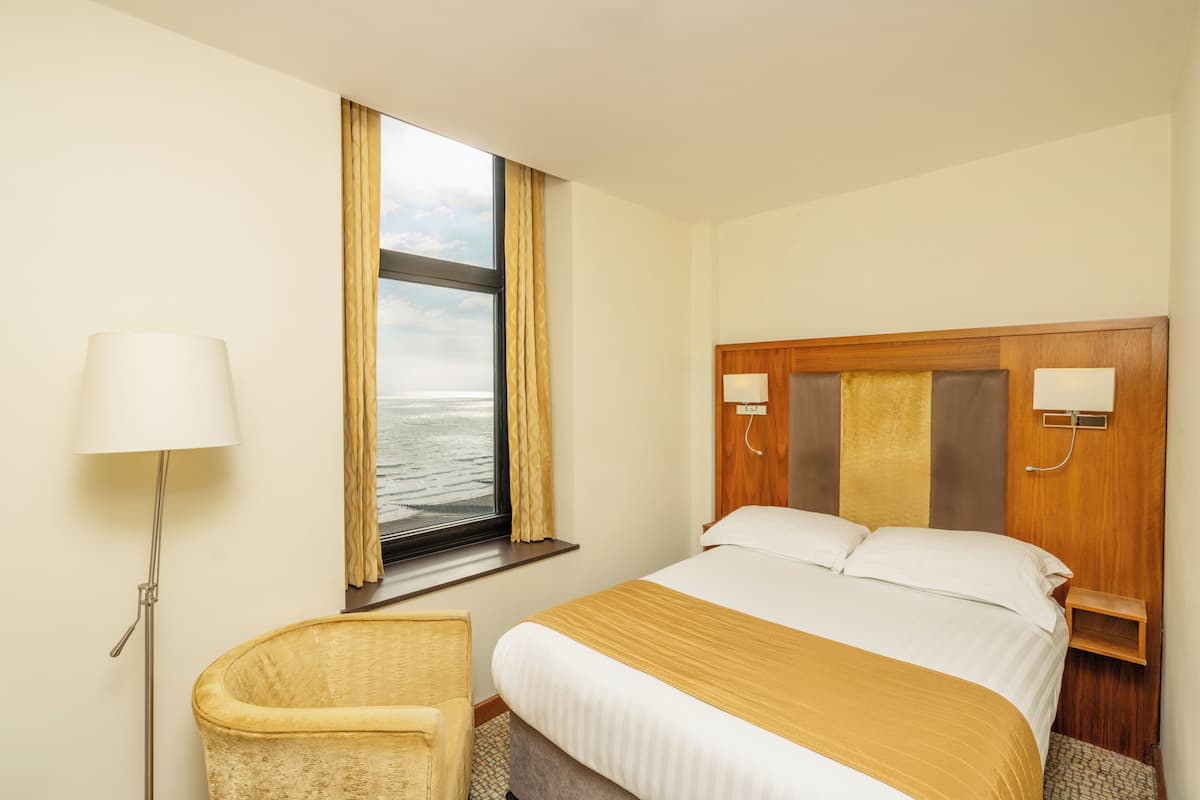 A hotel room with a bed, a chair, a lamp, a window with curtains, and a view of the water.