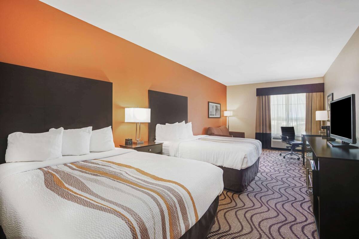 A hotel room with two beds, a desk, a TV, and a large window with curtains.