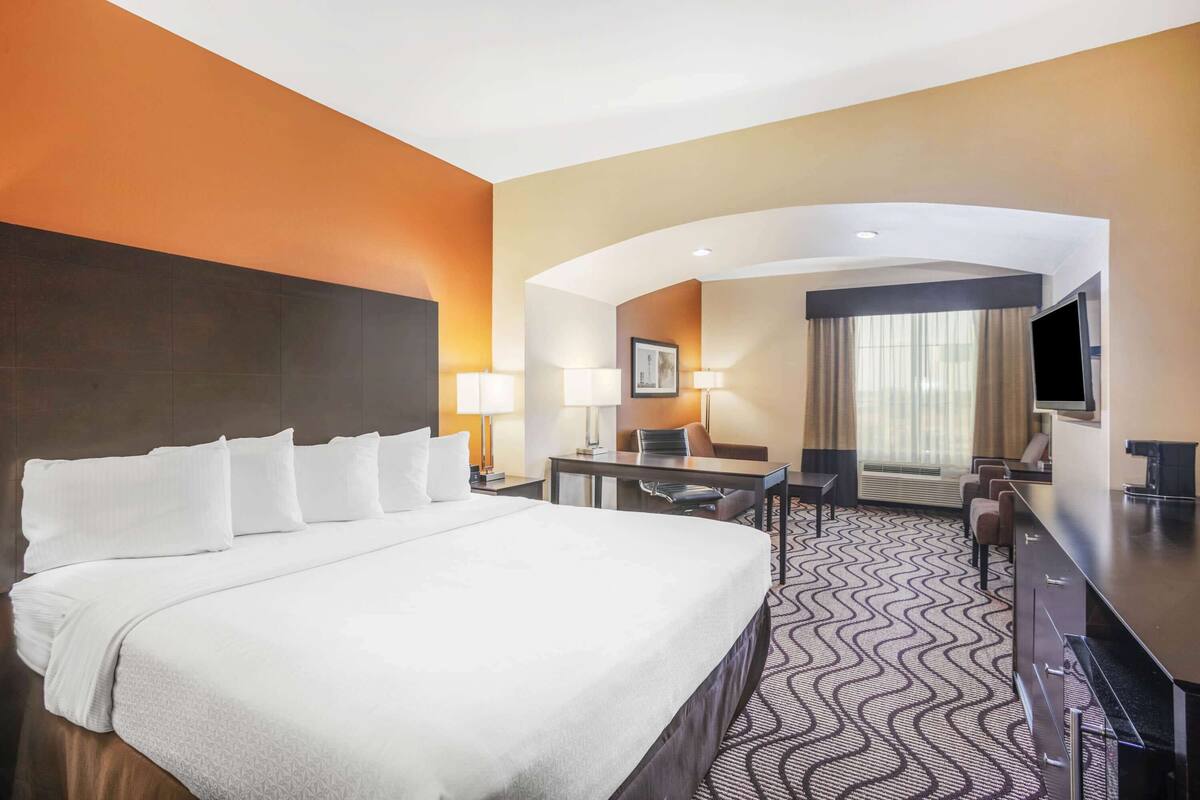 A hotel room with a large bed, a desk, two chairs, a TV, and a window with curtains.