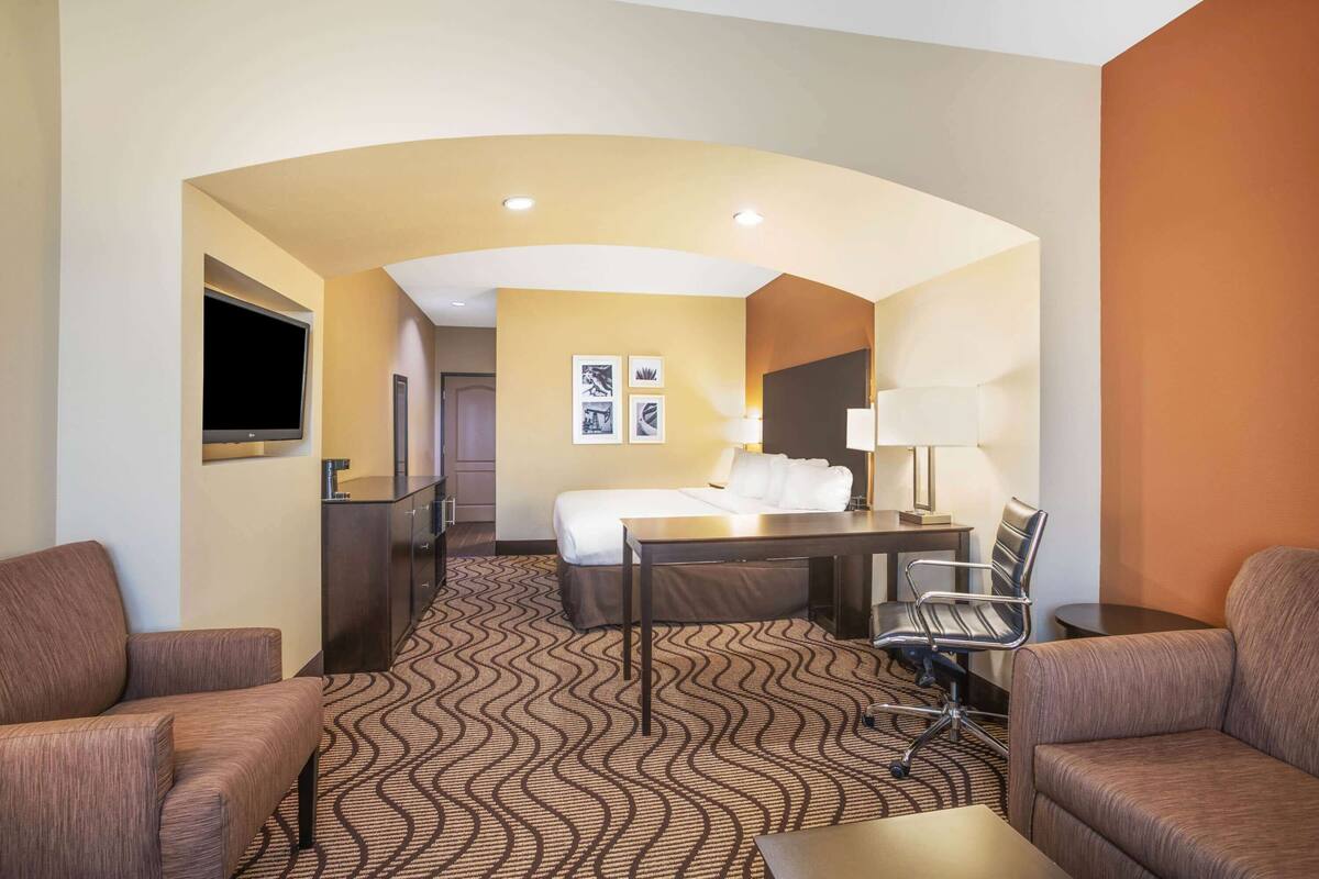 A hotel room with a bed, a desk, two armchairs, a TV, and a wall-mounted TV.