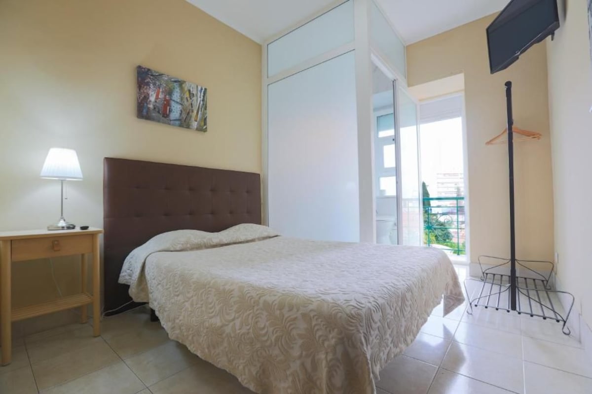 A bedroom with a bed, a nightstand, a lamp, a painting, a TV, and a balcony with a clothes rack.