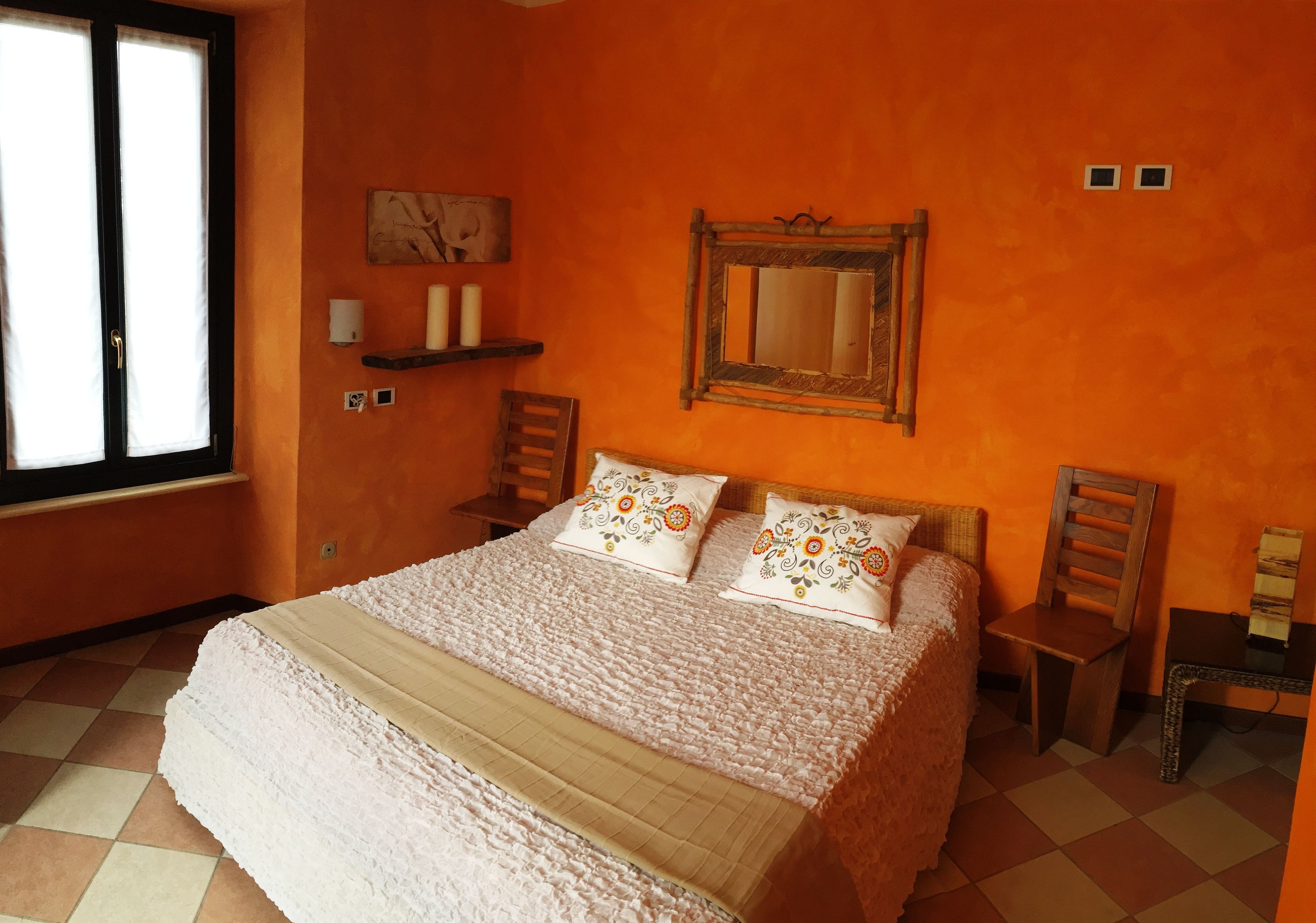 A bedroom with a large bed, two chairs, a small table, a window, and an orange wall.