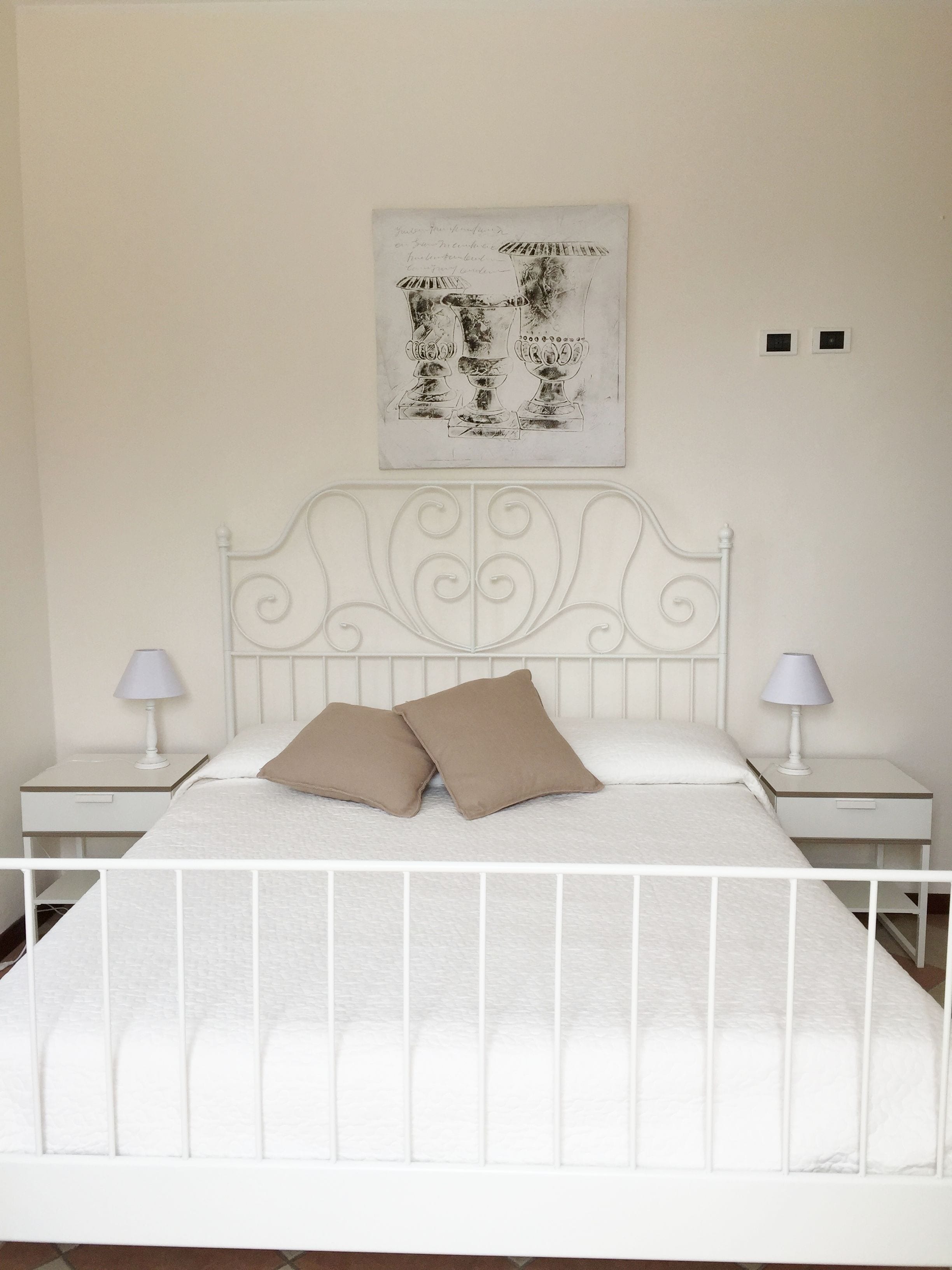 Une chambre à coucher avec un lit en métal blanc, deux tables de chevet avec des lampes, une tête de lit blanche et un tableau encadré au mur.