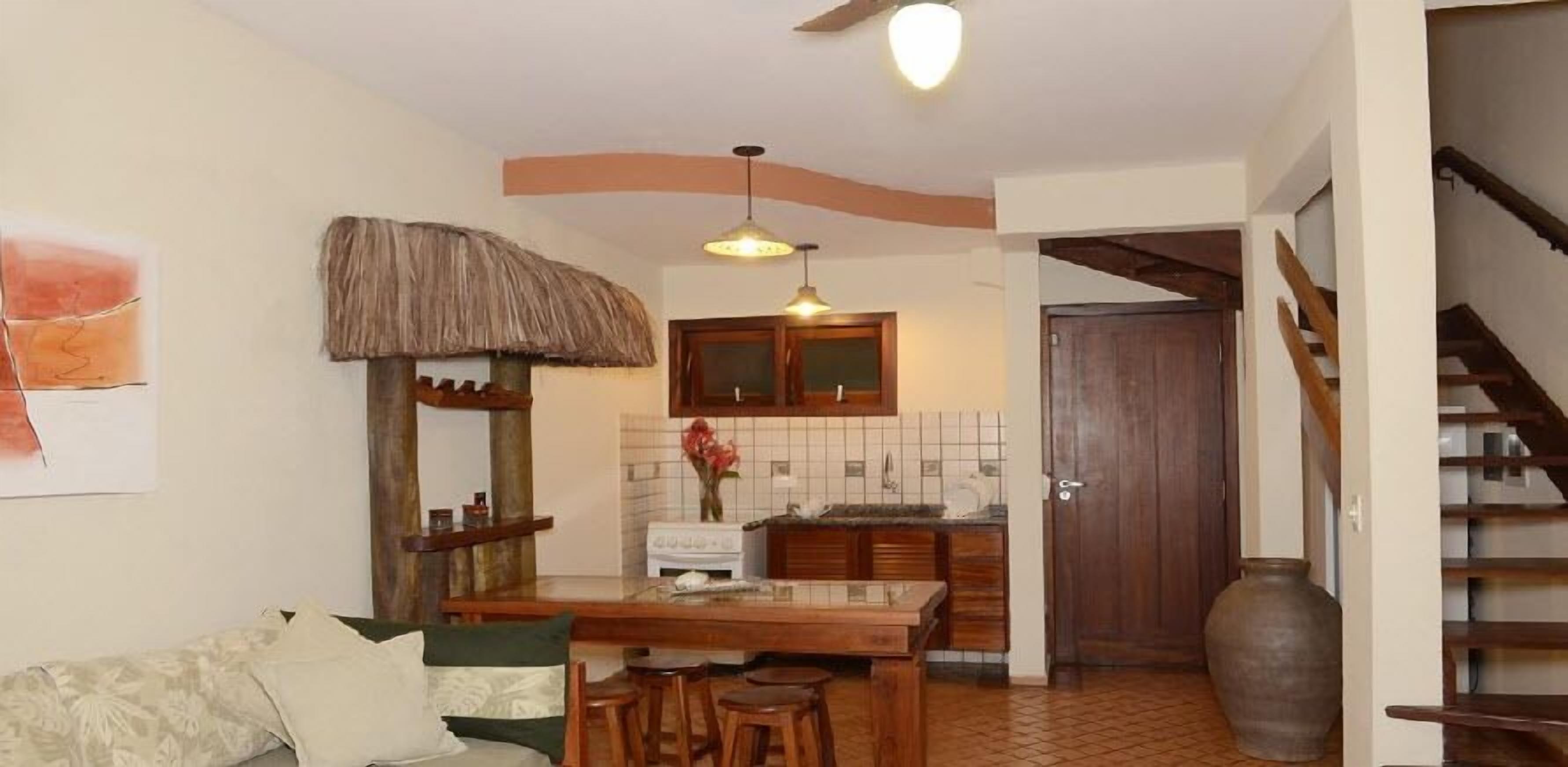 A compact kitchen with a stove, wooden stools, a hanging light fixture, and a rustic thatched ceiling.