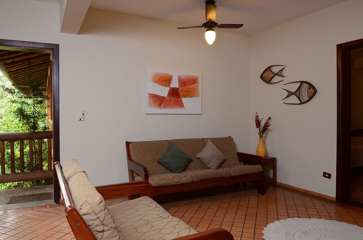 A room with a wooden sofa, a chair, a ceiling fan, a wall-mounted abstract painting, and a vase with flowers.