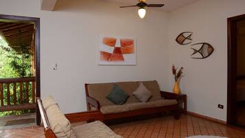 A room with a wooden sofa, a chair, a ceiling fan, a wall-mounted abstract painting, and a vase with flowers.