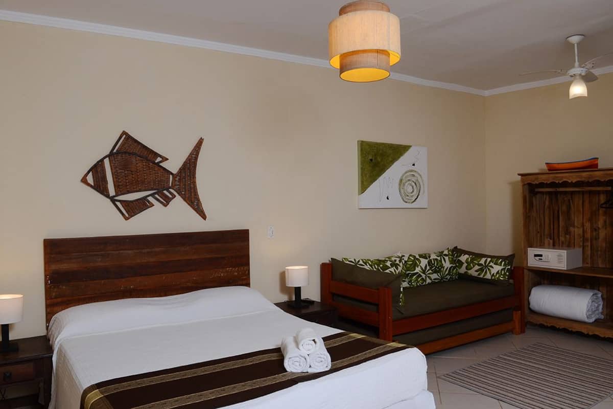 A hotel room with a bed, a wooden headboard, a wooden sofa, a wall art piece, a ceiling fan, and a small table with a lamp.