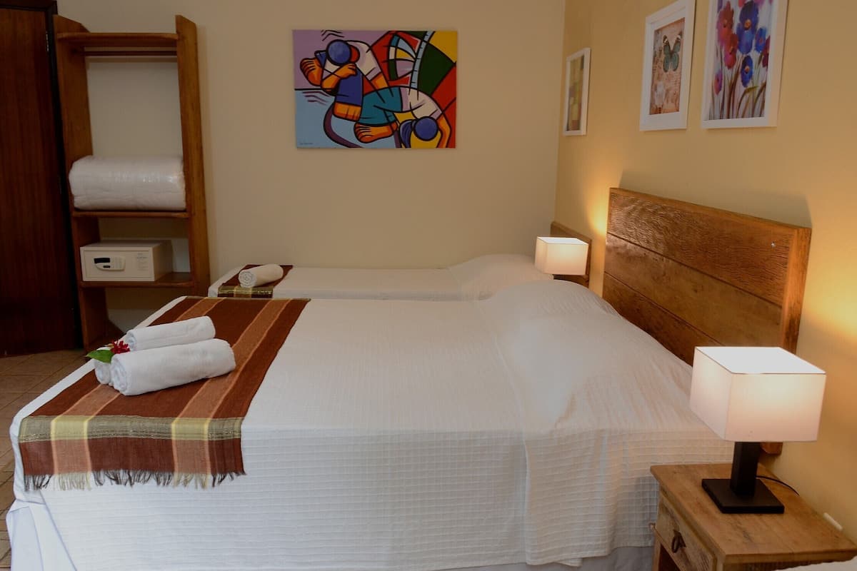 Una habitación de hotel con dos camas, un cabecero de madera, una mesita de noche con una lámpara, un armario y un cuadro en la pared.