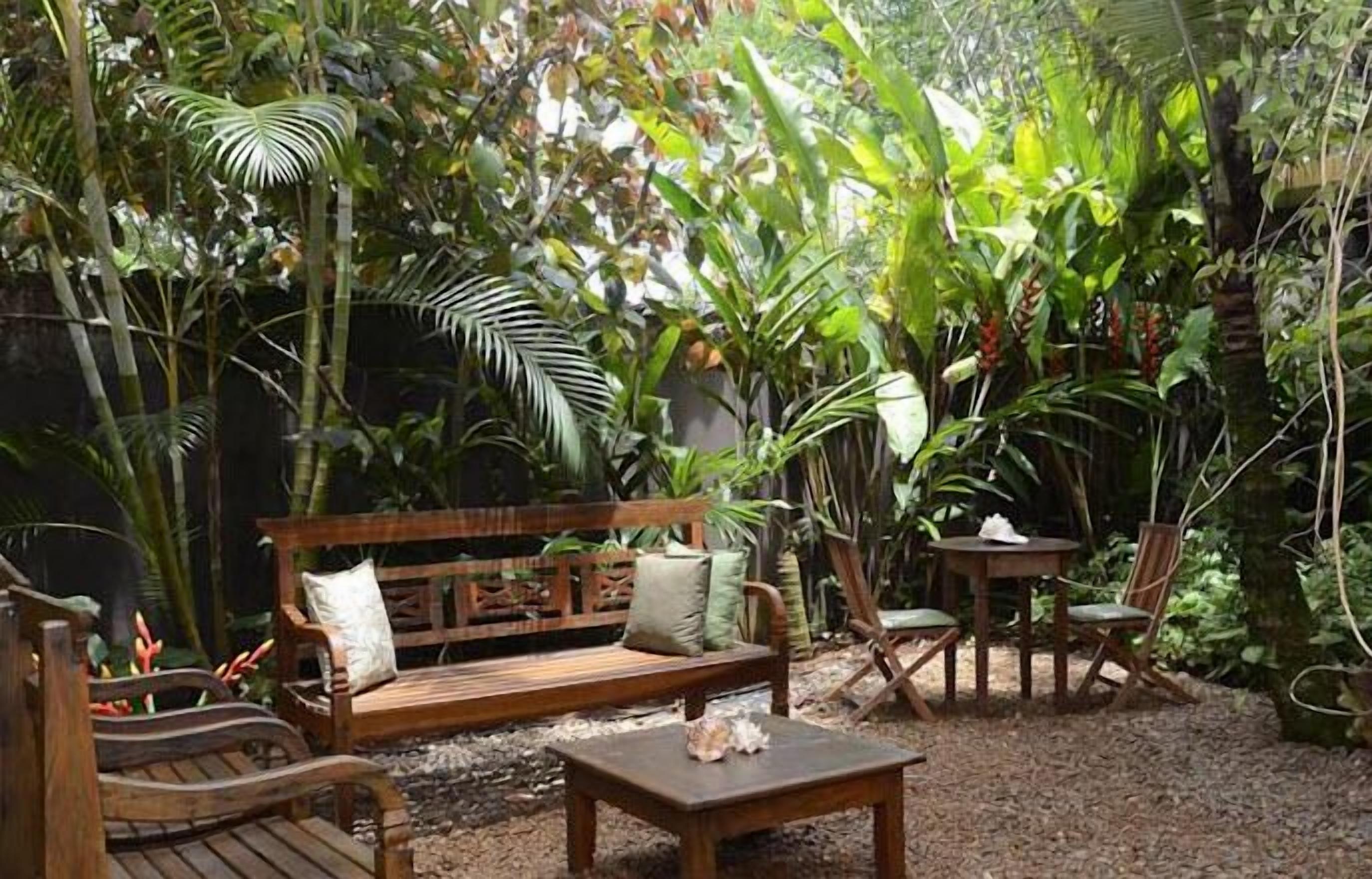 Una panca di legno, un tavolino e sedie in un giardino con vegetazione lussureggiante e pavimento in pietra.