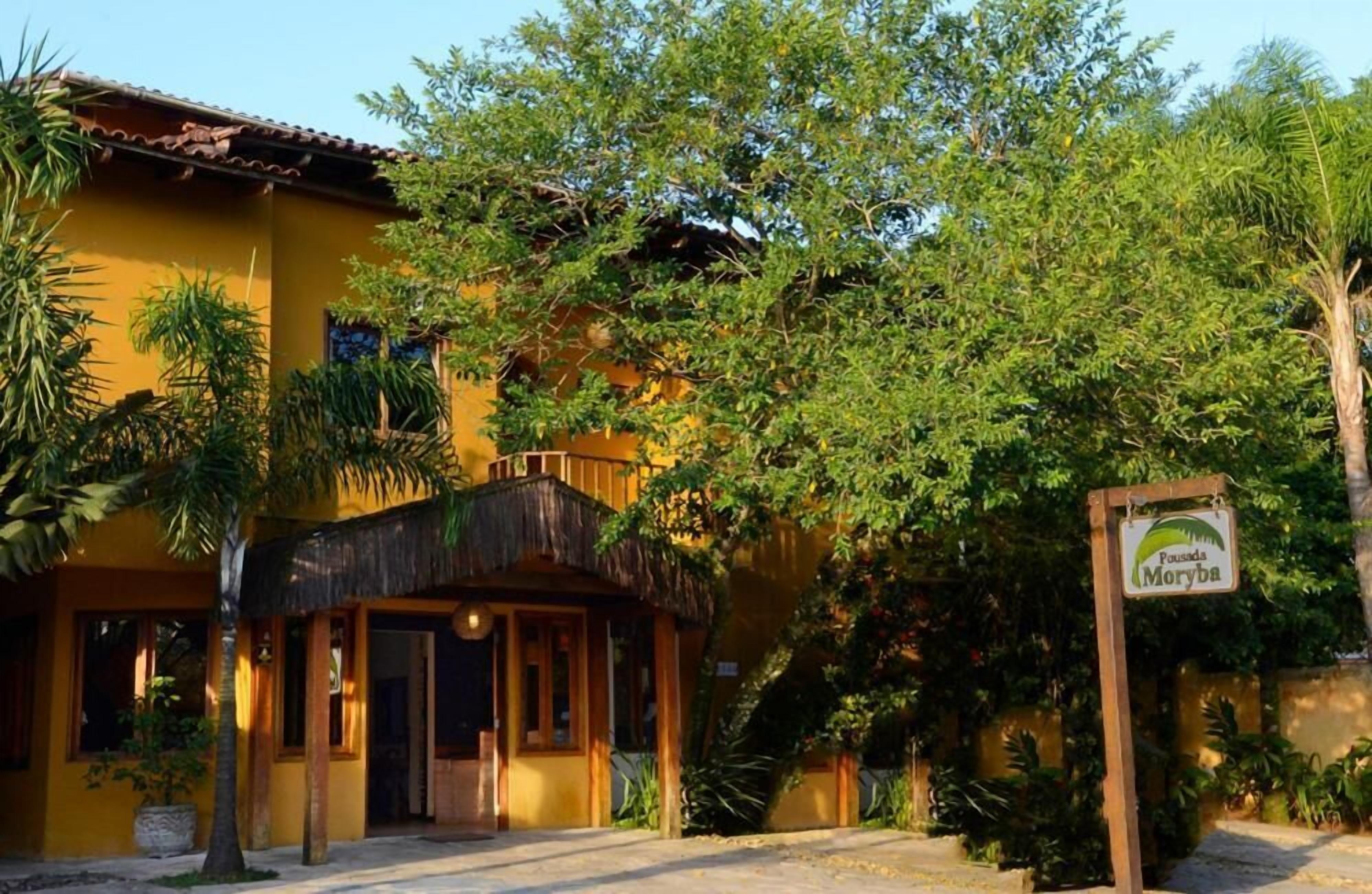 A yellow building with a thatched-roof structure in front, surrounded by lush greenery.