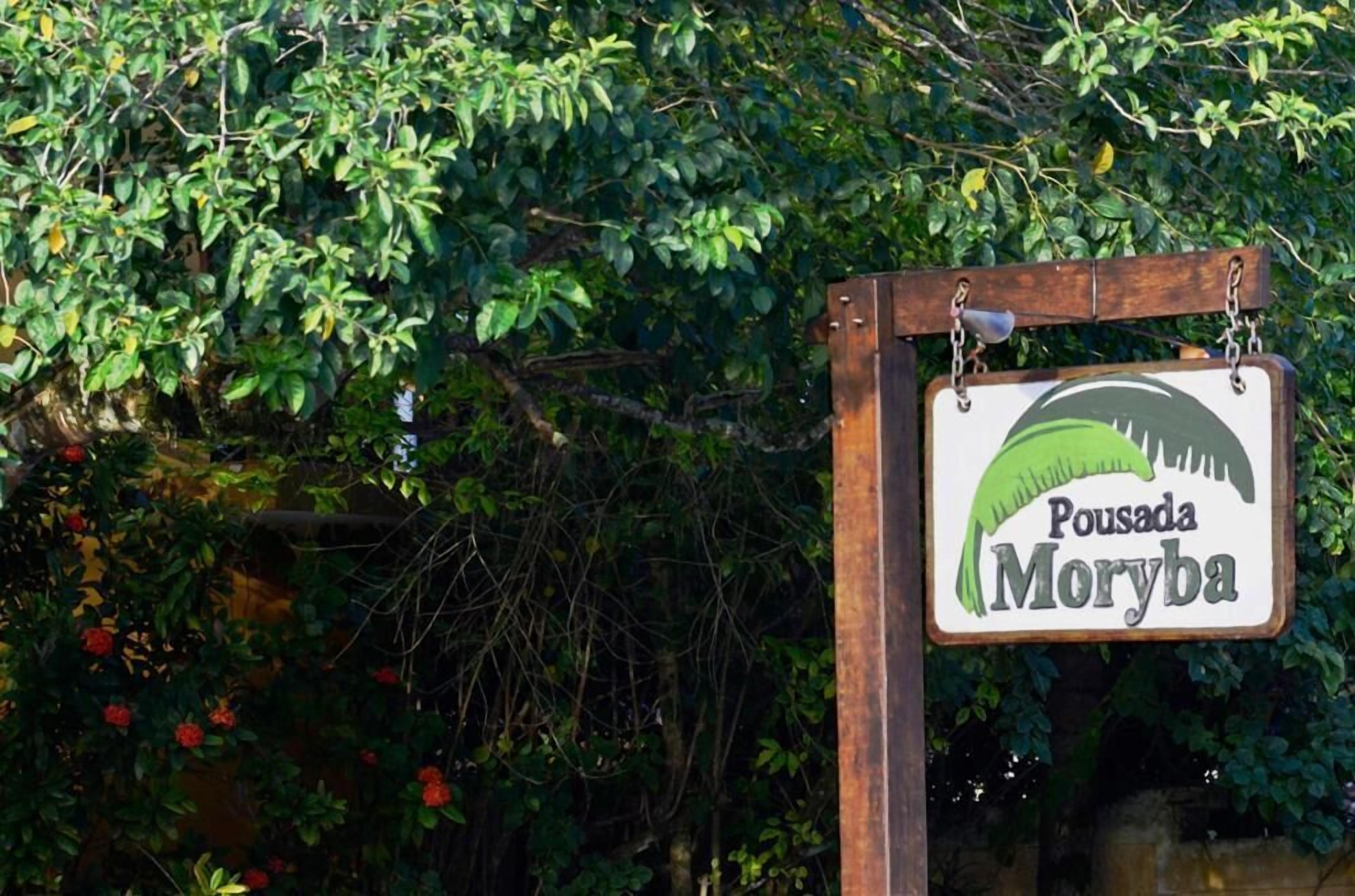 Ein Schild mit dem Schriftzug 'Pousada Moryba' vor üppigem Grün und einer Steinmauer.