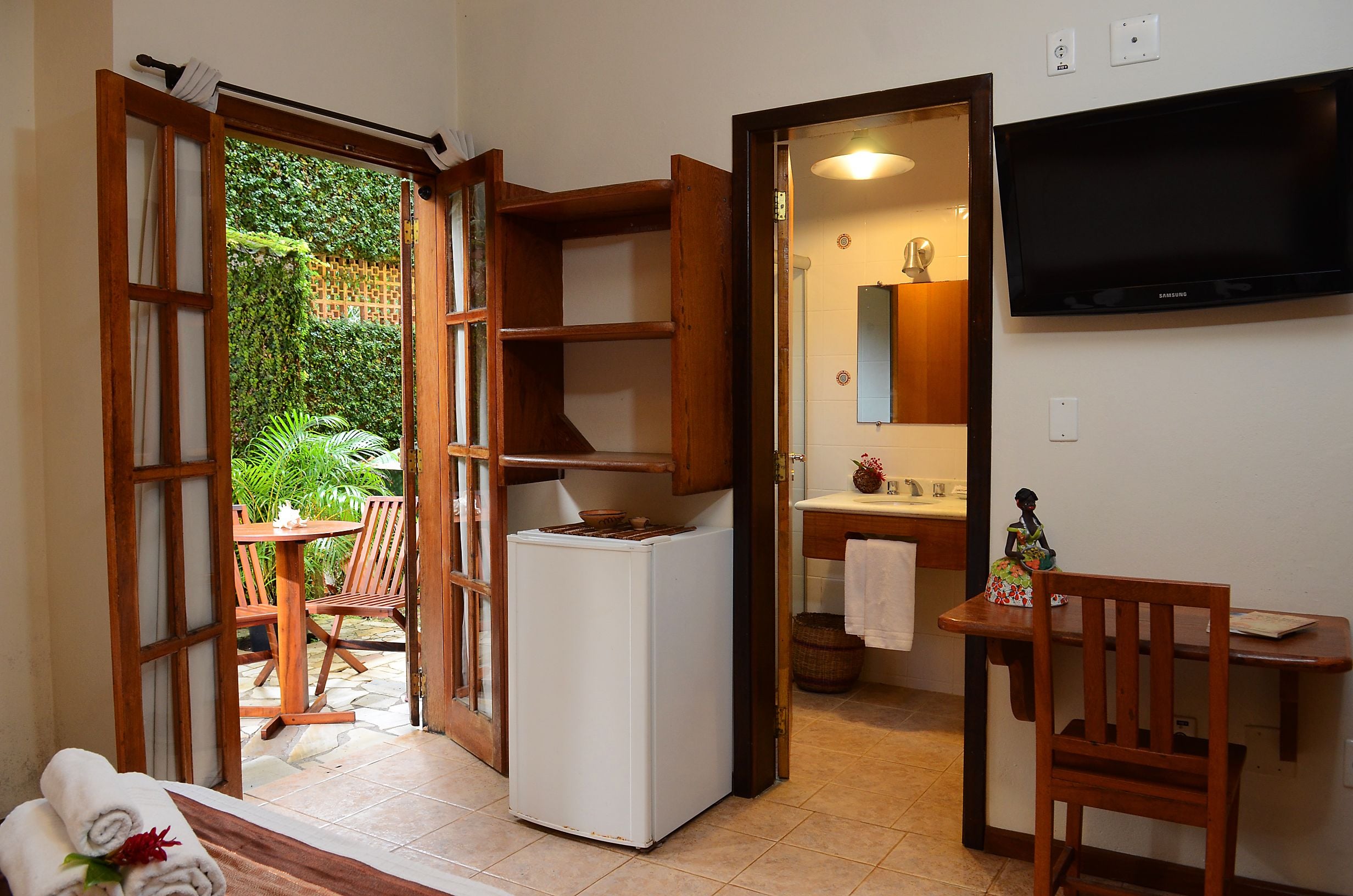 A compact room with a refrigerator, a wooden chair, a small table, and a television mounted on the wall.
