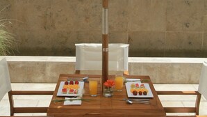 Una mesa de madera con platos de fruta y jugo, con una piscina y muros de piedra de fondo.