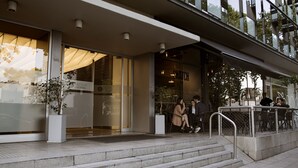 Una entrada de hotel moderna con fachada de vidrio, área de estar al aire libre y una escalera que lleva a la entrada.