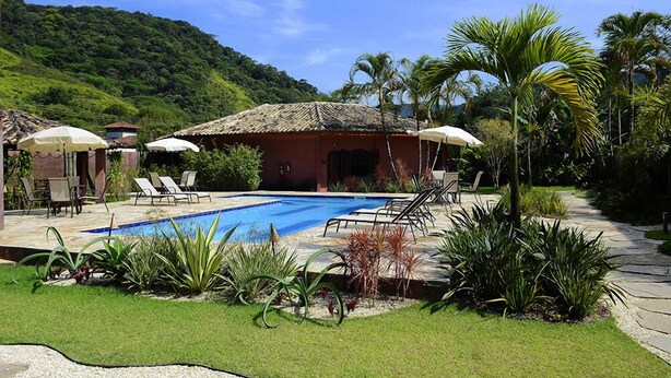 A pool area with lounge chairs and umbrellas, surrounded by tropical plants and a mountain backdrop. - Pousada Tupinambá (São Sebastião)