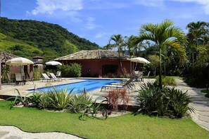 Zona de piscina con sillones de descanso y sombrillas, rodeada de plantas tropicales y una montaña al fondo.