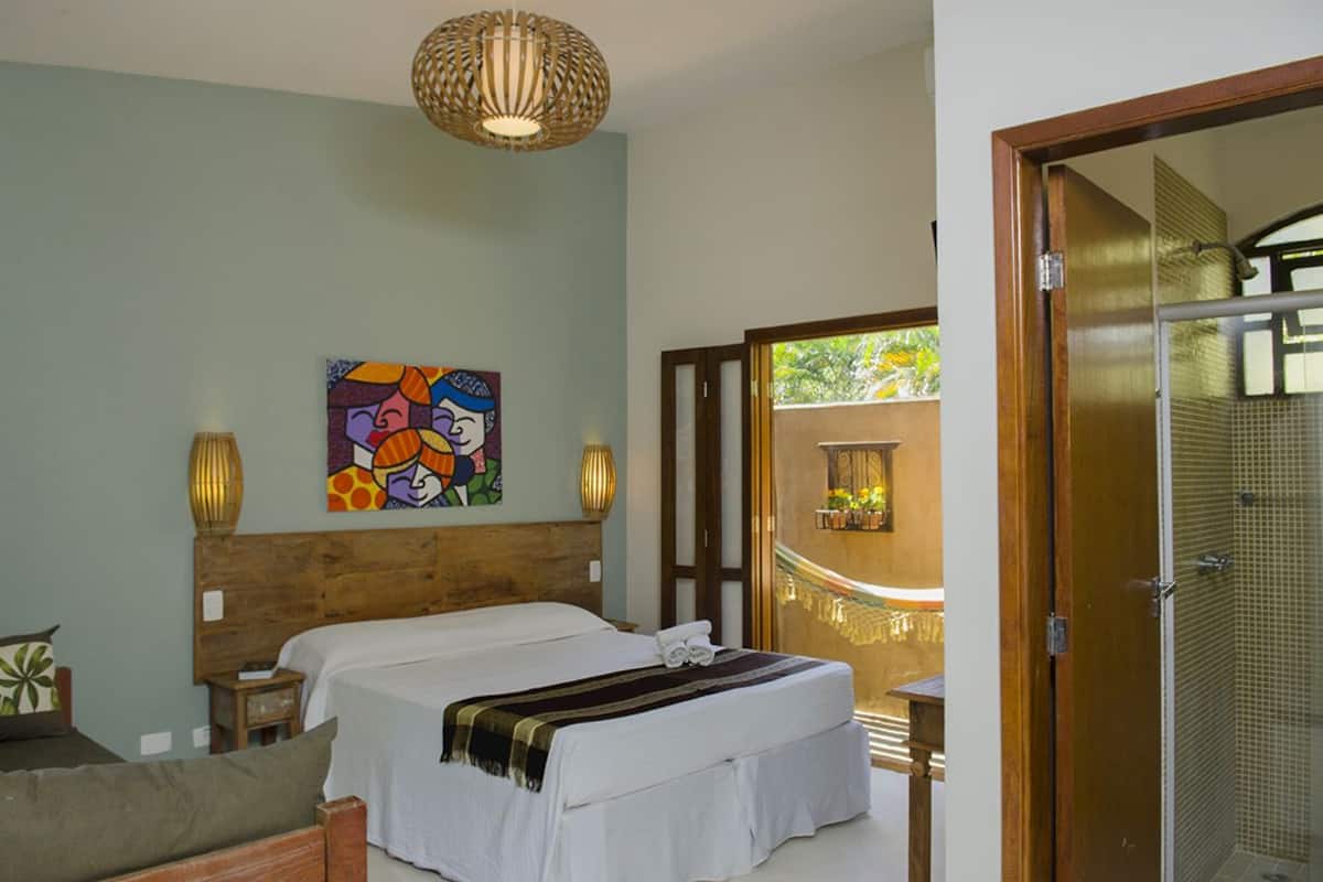 A bedroom with a wooden headboard, a bed with white linens, a colorful abstract painting, a small bedside table with a lamp, and a view of a balcony with a railing.