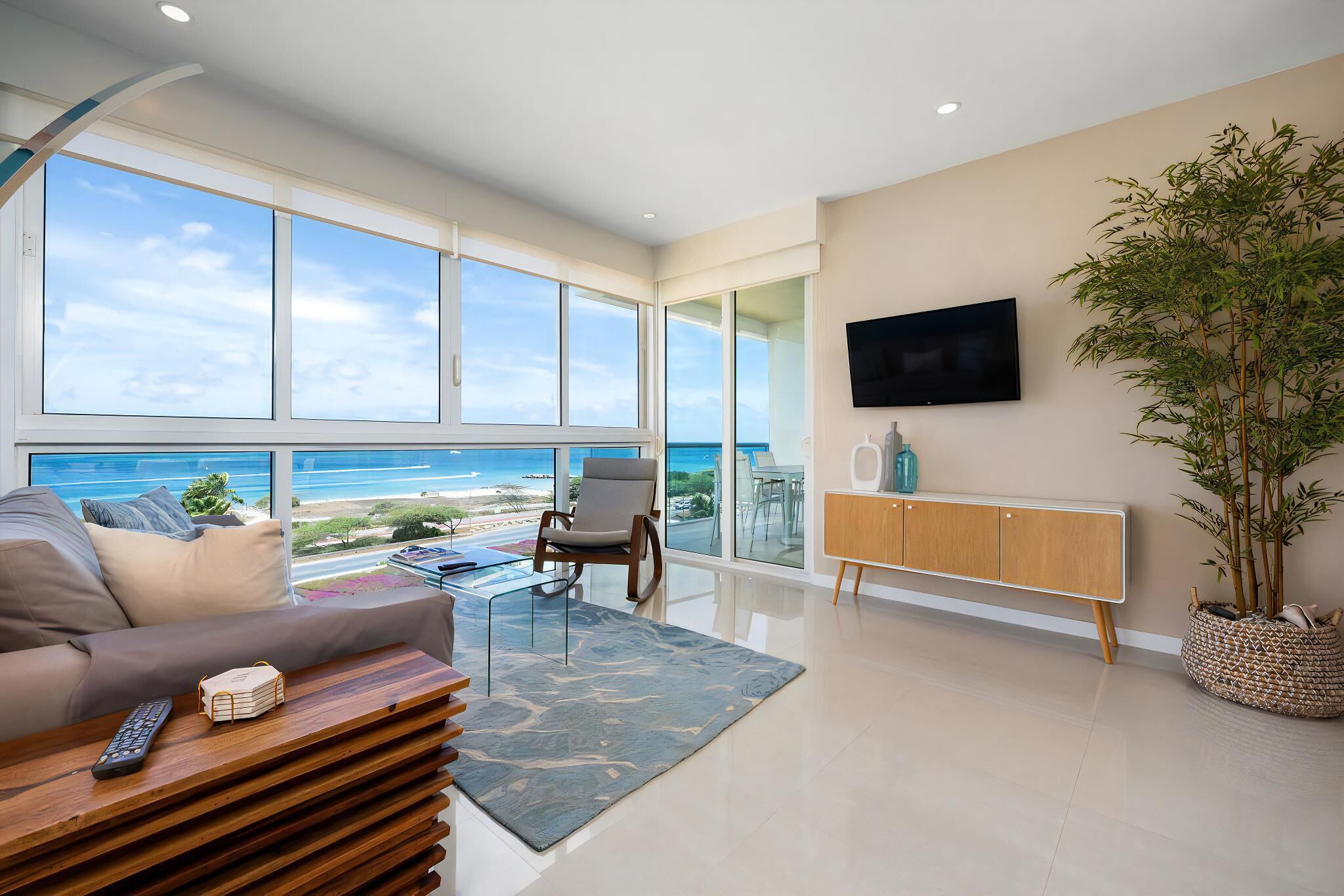 A modern living room with a large window overlooking the ocean, a sofa, a coffee table, a TV, and a potted plant.