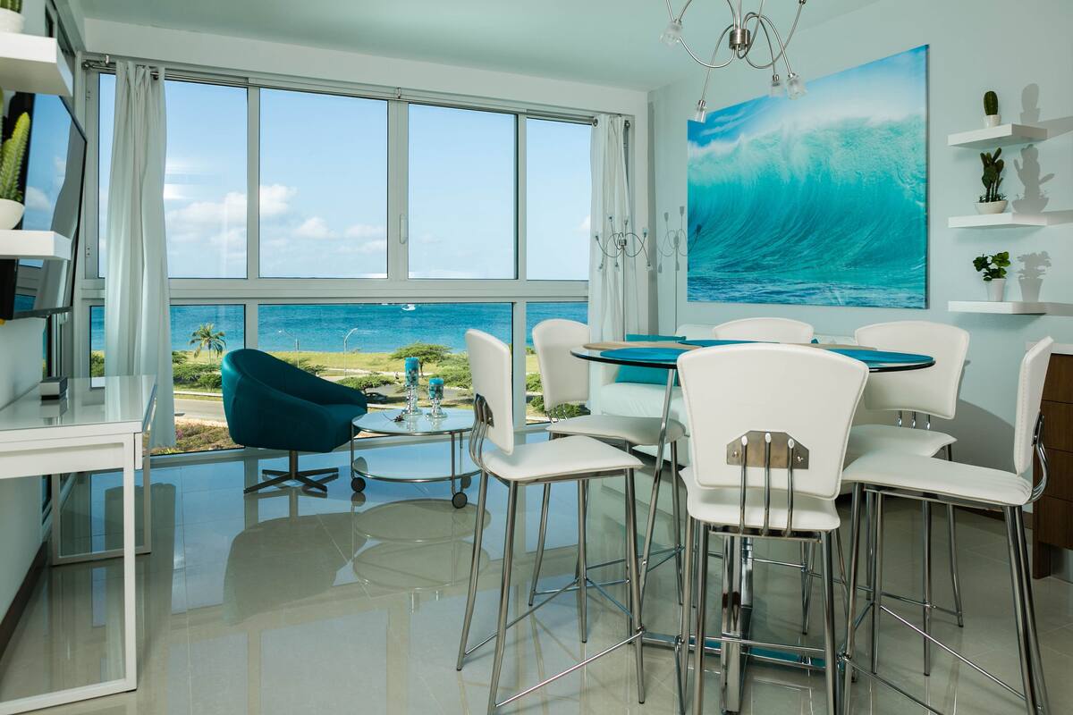 A modern dining area with a view of the ocean, white chairs, a glass table, and a large wave painting on the wall.