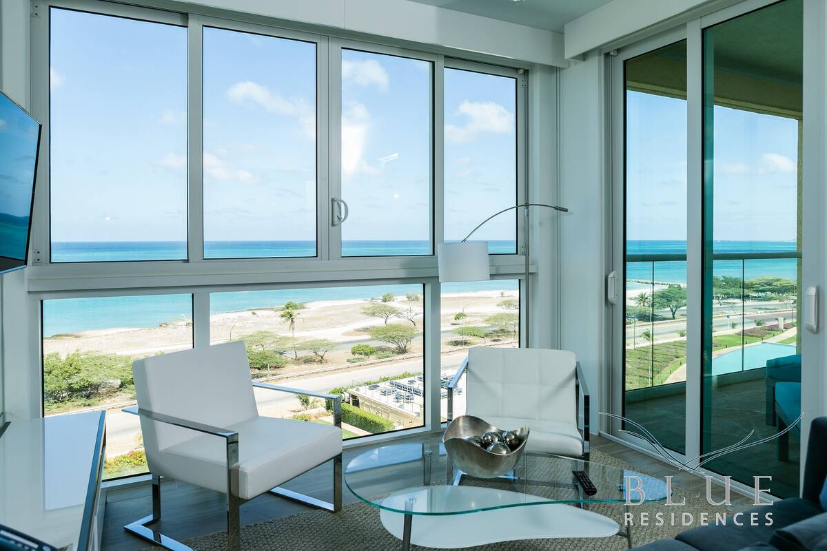 A modern living room with large windows, a glass coffee table, and two white chairs.