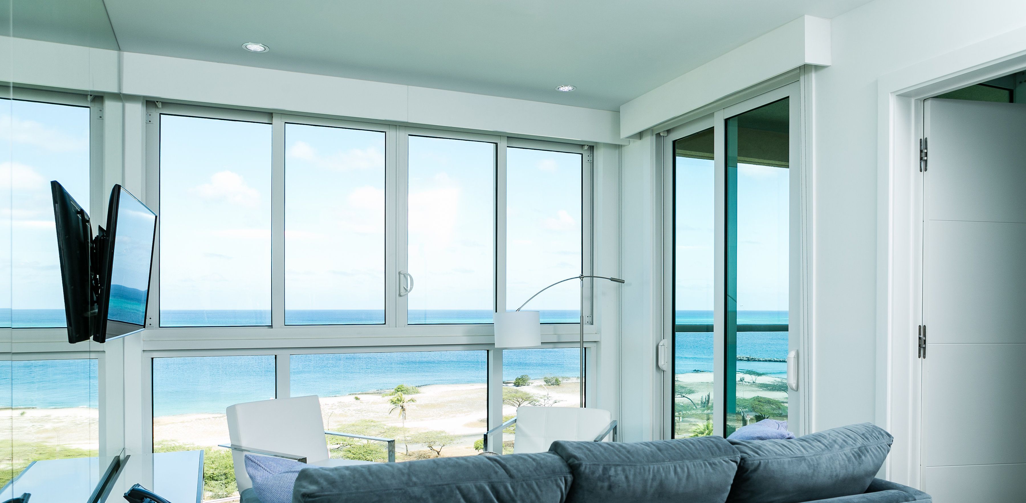 A modern room with large windows overlooking a beach, a flat-screen TV mounted on the wall, and a grey sofa.