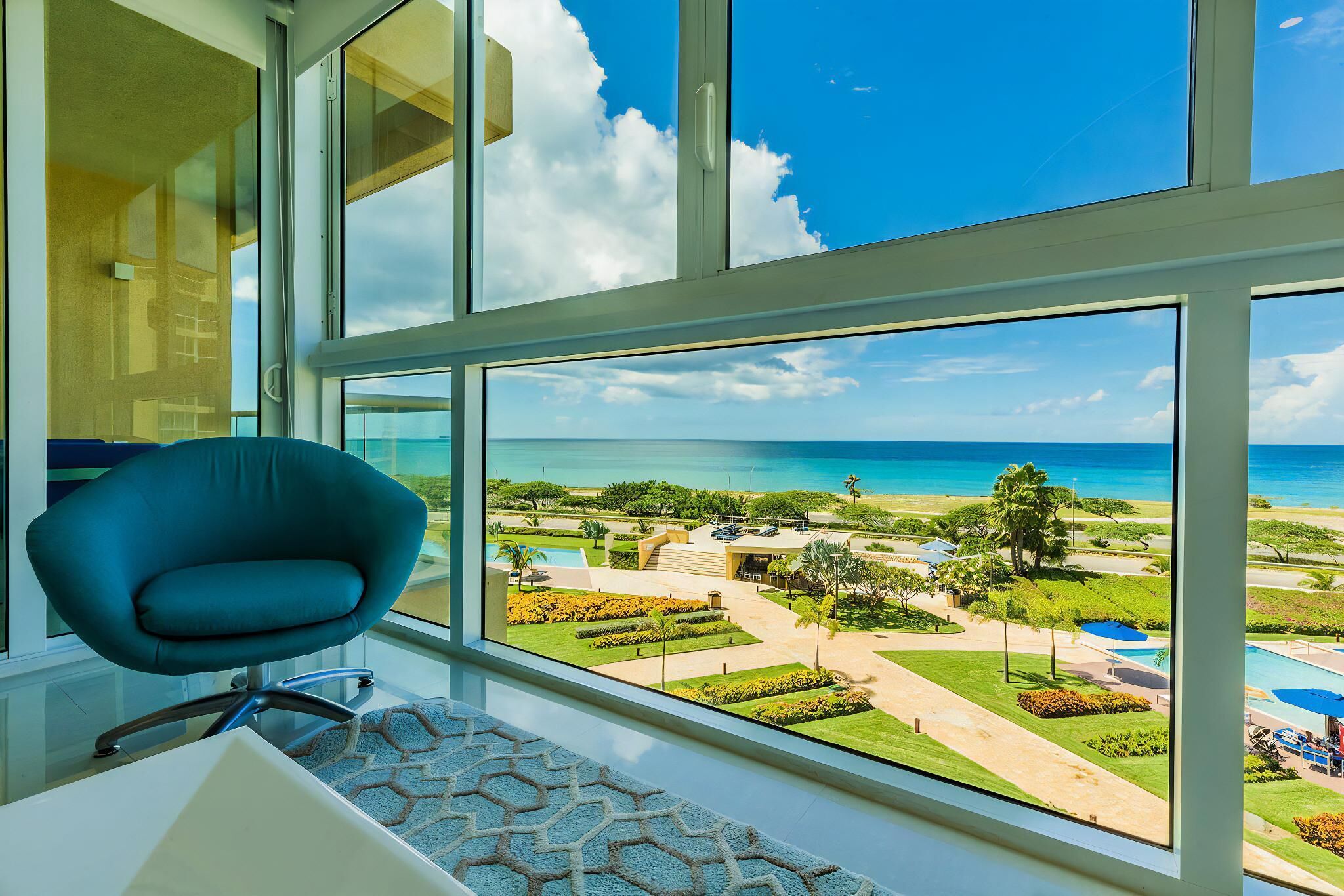 A room with a large window overlooking a landscaped area with a pool and a view of the ocean.