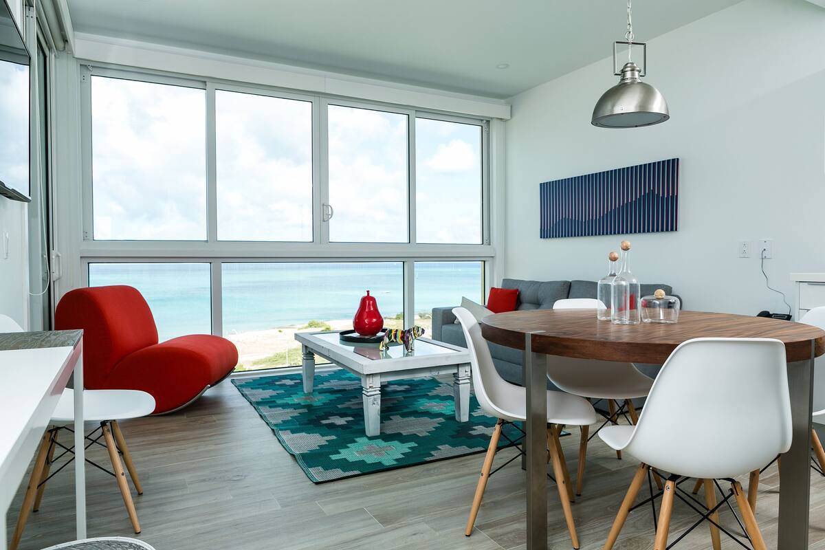 A modern living room with a large window, a dining table, and a view of the ocean.