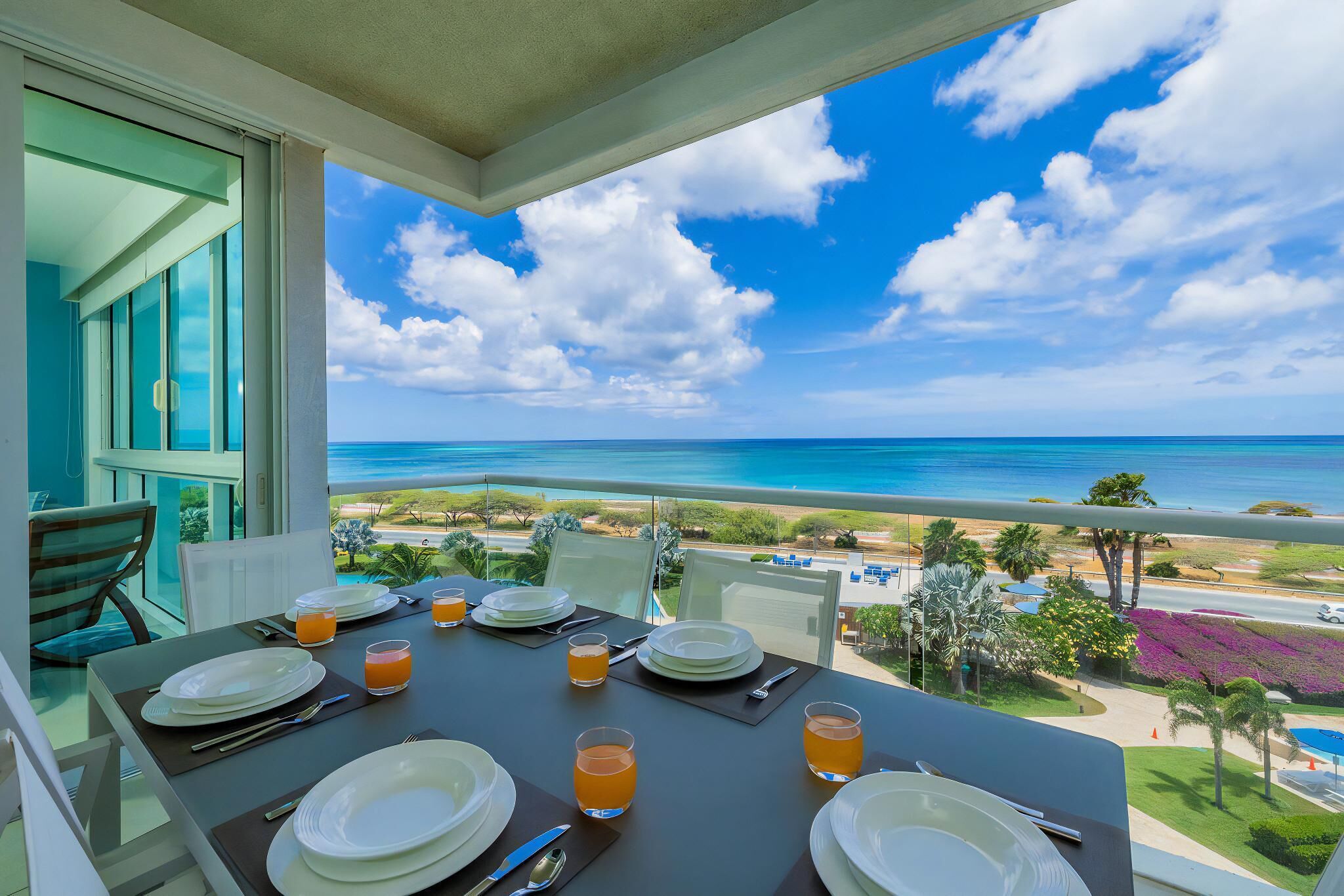 Ein Esstisch für vier Personen ist gedeckt mit Tellern, Besteck und Gläsern Orangensaft, mit Blick auf Strand und Meer.