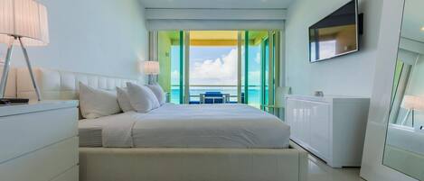 A modern hotel room with a large bed, a TV mounted on the wall, and a view of the sea through large windows.