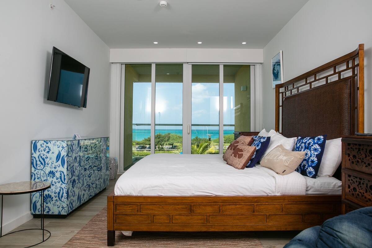 A bedroom with a large bed, a TV mounted on the wall, a glass door leading to a balcony with a view of the ocean, and a blue patterned cabinet.