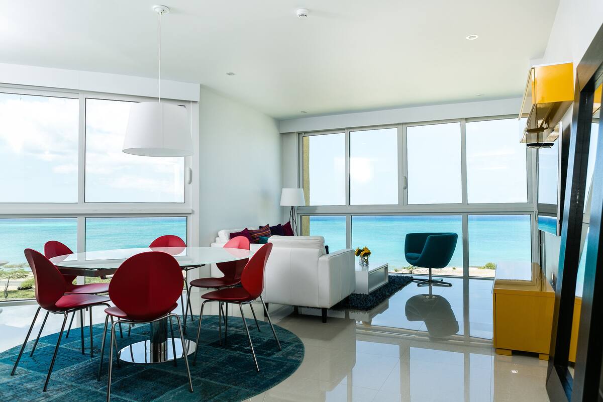 A modern room with a large window overlooking the ocean, a white sofa, red chairs, and a dining table.