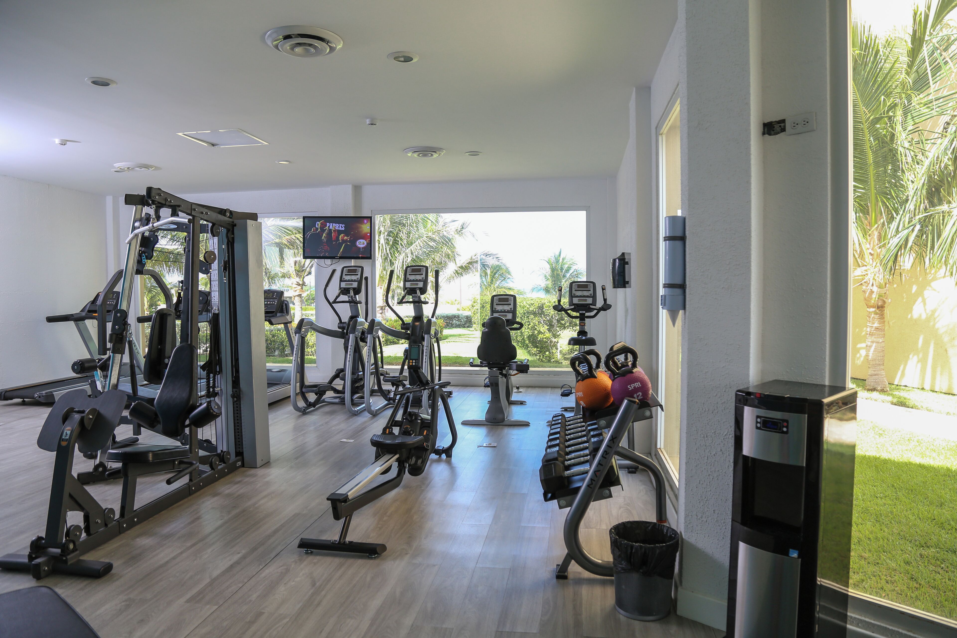 Un gimnasio con equipos de ejercicio variados, un dispensador de agua y vistas a palmeras afuera.