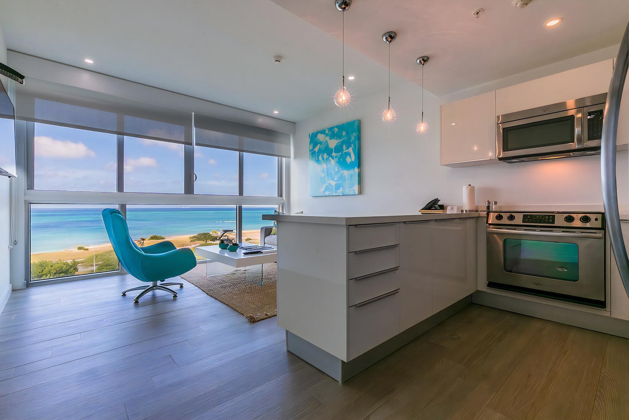 Uma cozinha moderna com uma janela ampla com vista para a praia, uma cadeira verde-água e um forno de aço inoxidável.